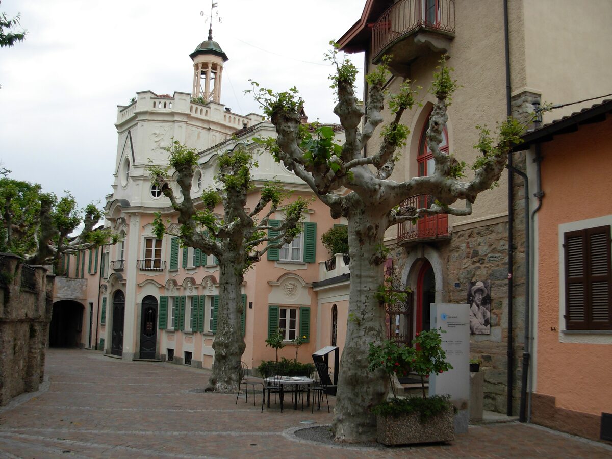 A pale tower house and pollarded plane trees on a cobbled courtyard in Montagnola, next to the Hermann Hesse Museum