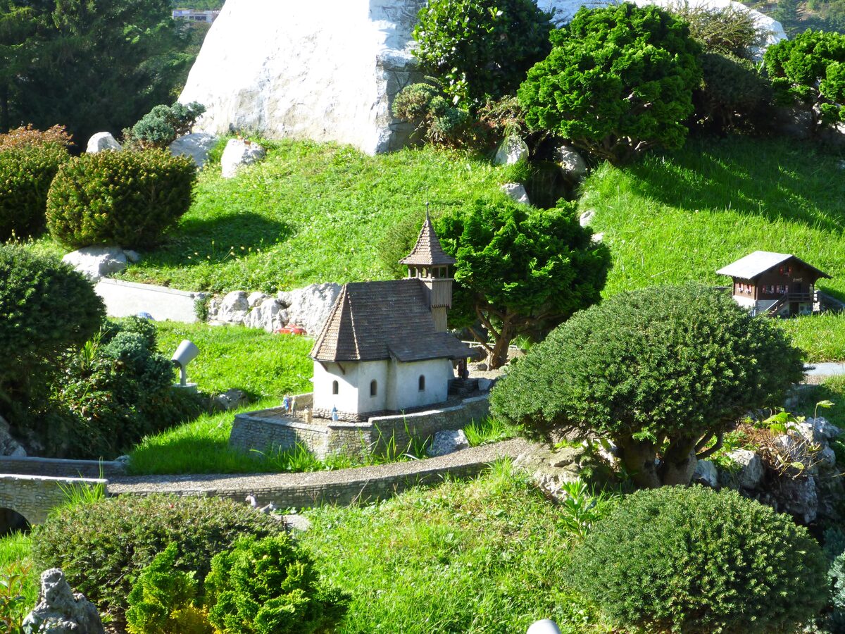 A miniature stone chapel and clipped trees on a green slope inside the Swissminiatur park at Melide