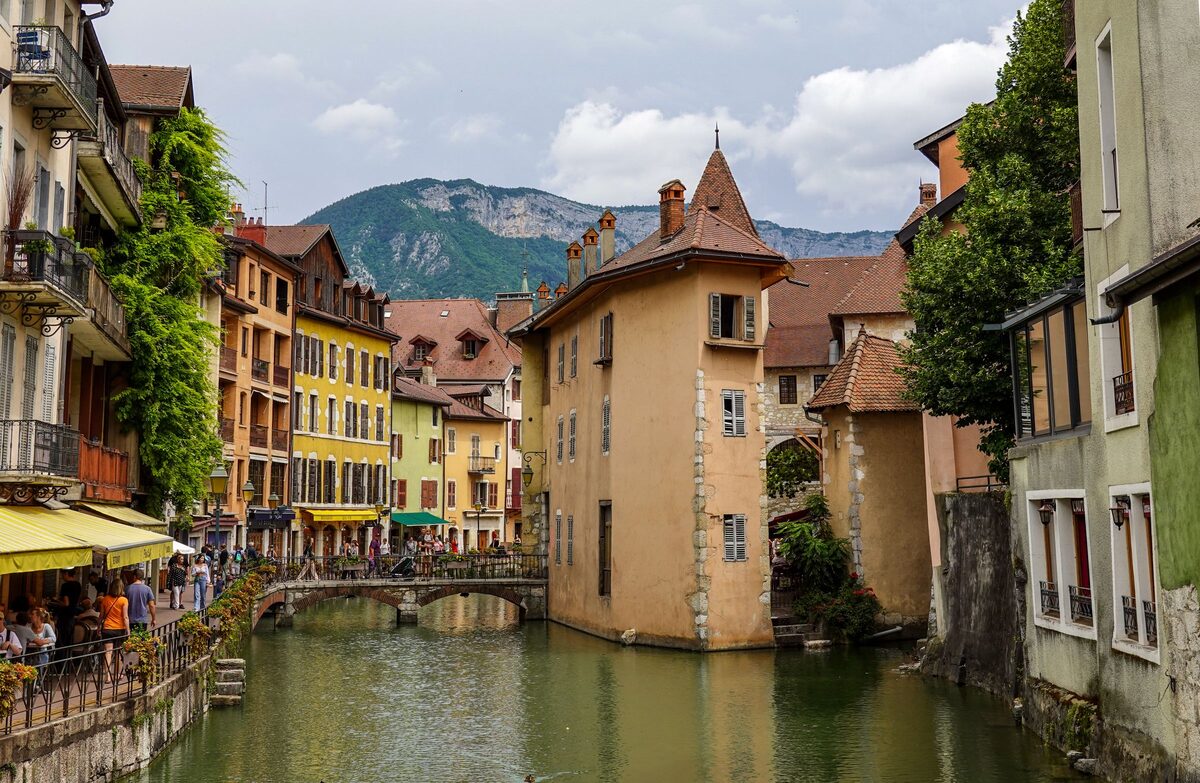 A canal in the old town of Annecy lined with pastel houses and flower boxes, with a turreted old prison on an island and mountains behind