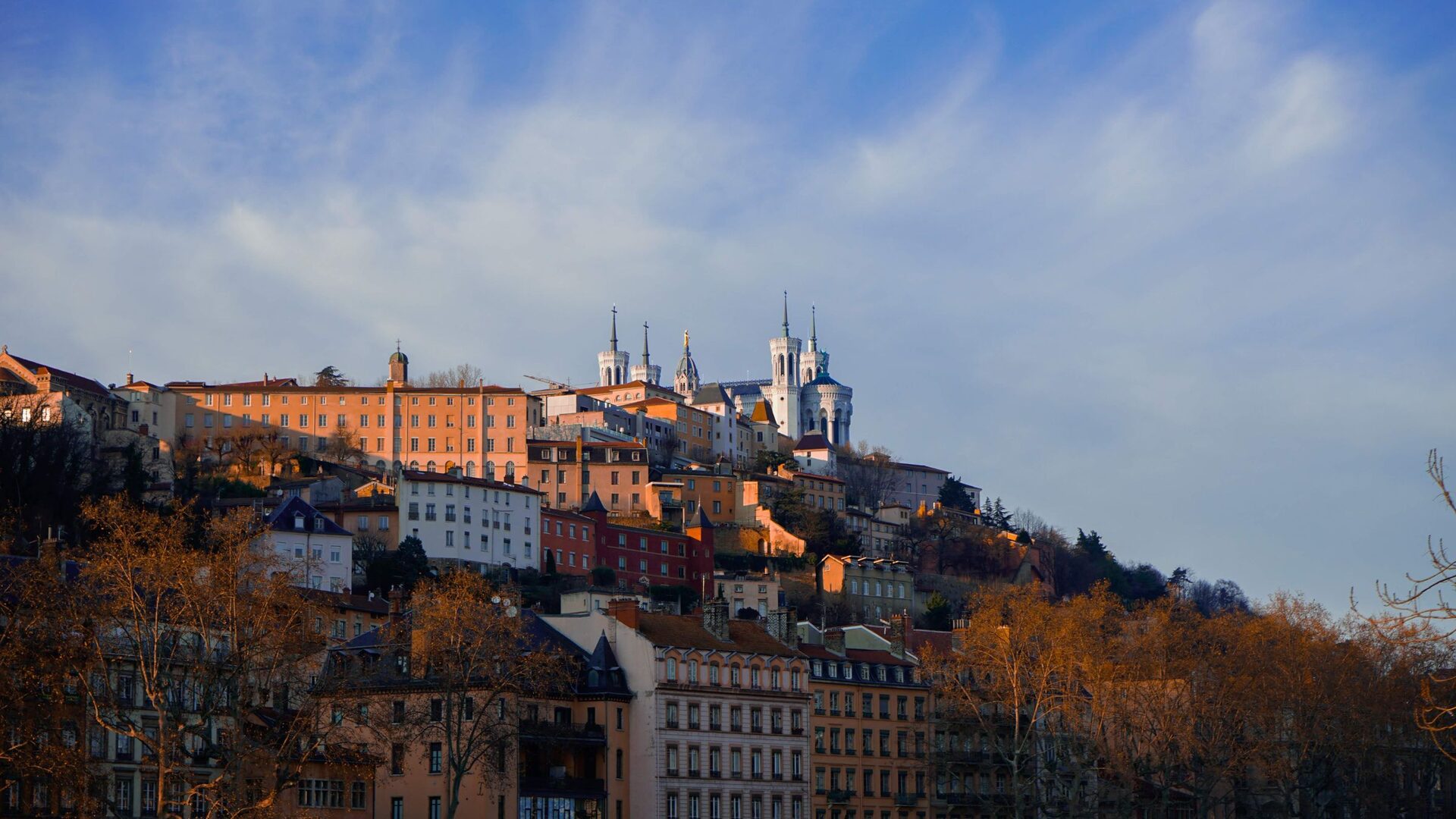 Fourvière hill in Lyon in warm evening light, with the white basilica on top above old ocher and pink houses and bare autumn trees