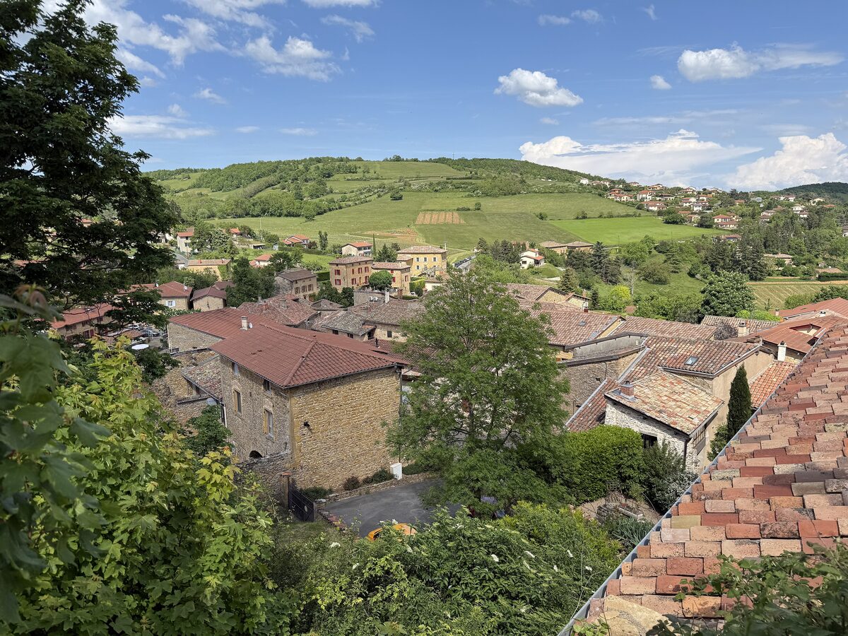 The tiled roofs of Oingt with green hills and vineyards rolling away behind under a blue sky with small clouds
