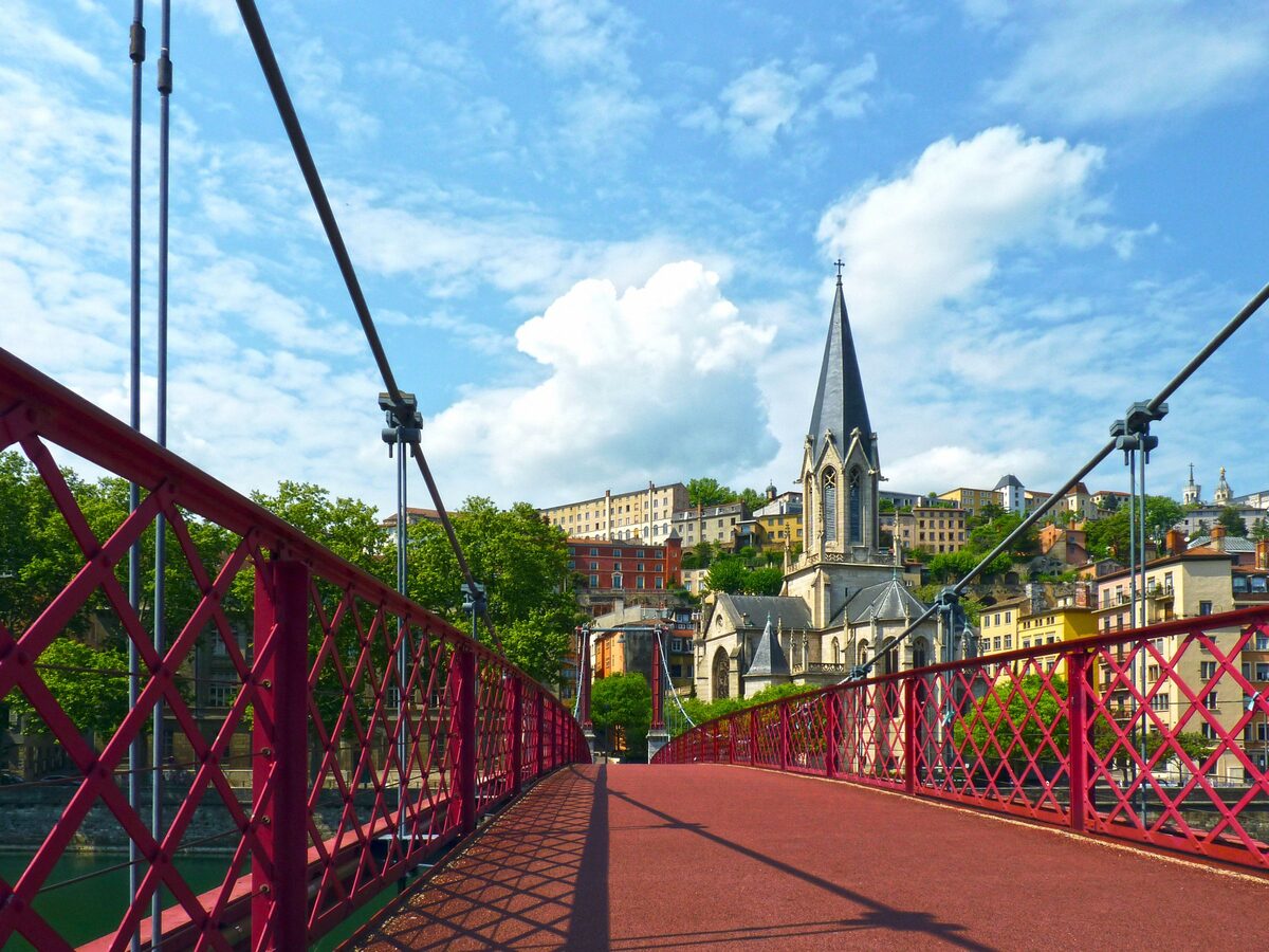 The red Saint-Georges footbridge over the Saône leading to the Gothic spire of the church of Saint-Georges, under a blue sky with white clouds