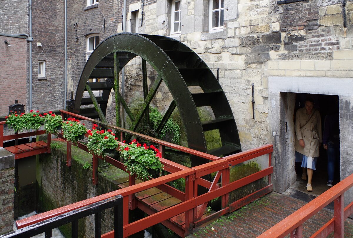 The big wooden waterwheel of the Bisschopsmolen turning beside an old stone building, with red railings and geraniums in the foreground