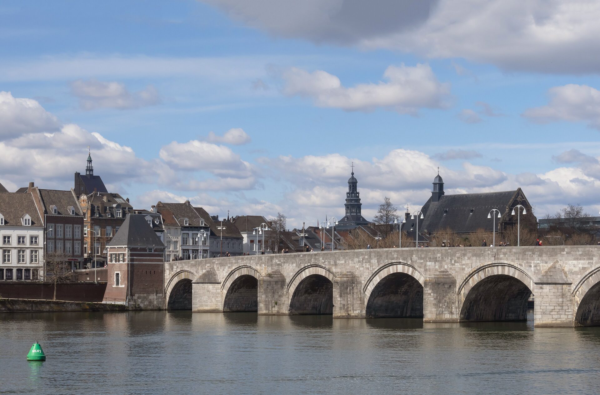 The stone arches of the Sint Servaasbrug over the Maas in Maastricht, with old houses and church towers behind under a blue sky with white clouds