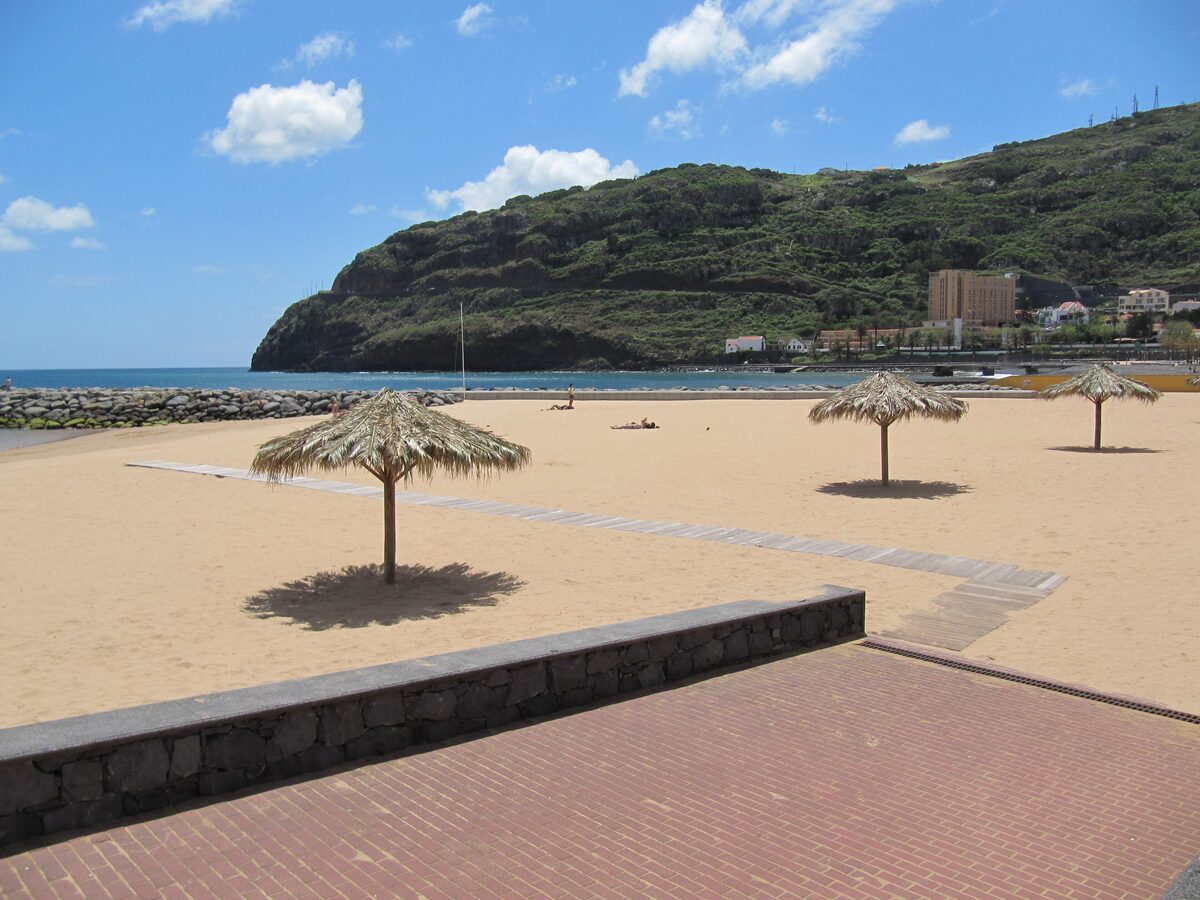 The golden sand beach at Machico on a sunny day, with thatched parasols, a breakwater and a green headland behind