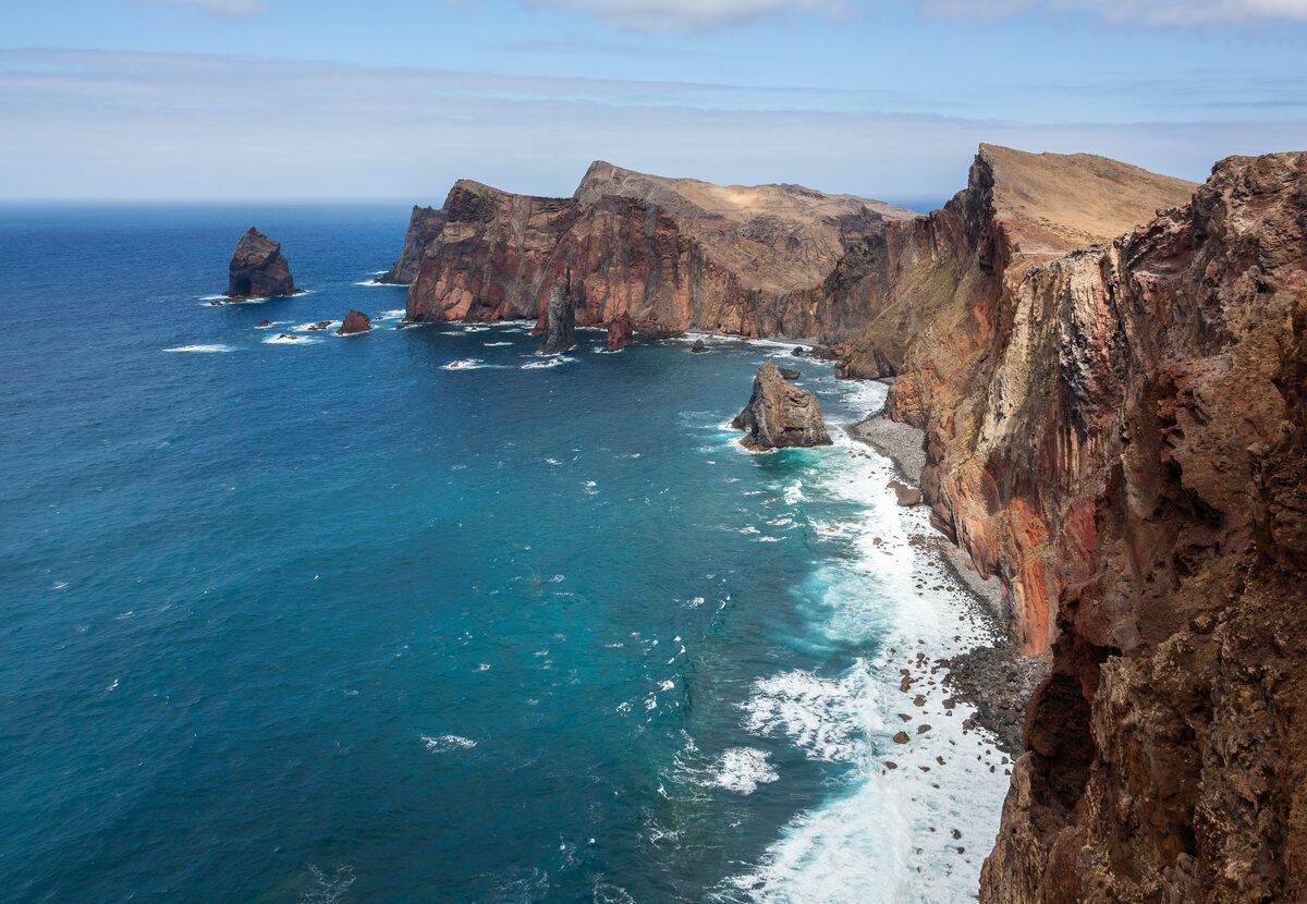 Red and brown cliffs with sea stacks at Ponta de São Lourenço, with white surf breaking on the dark blue sea below