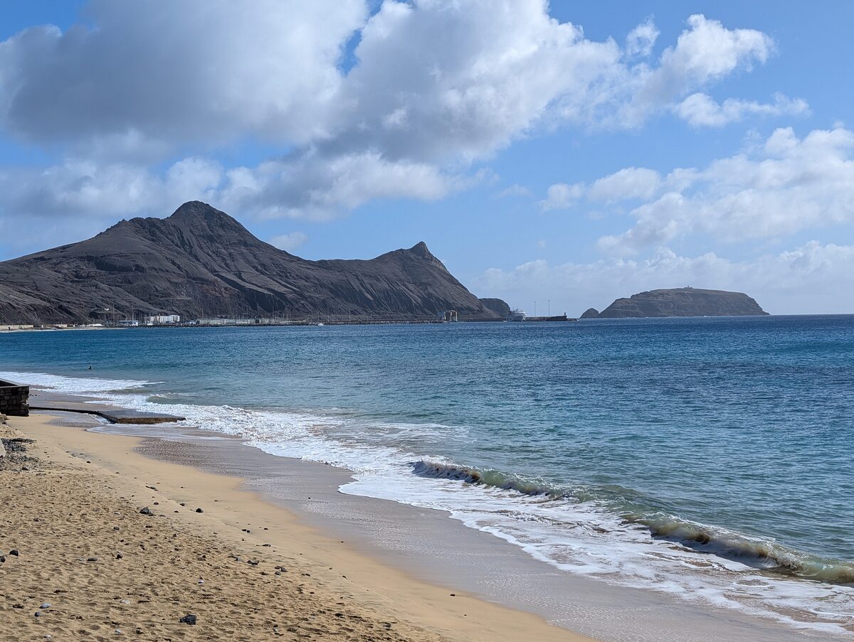 Waves on a sandy Porto Santo beach, with dark volcanic hills and a small islet across the bay