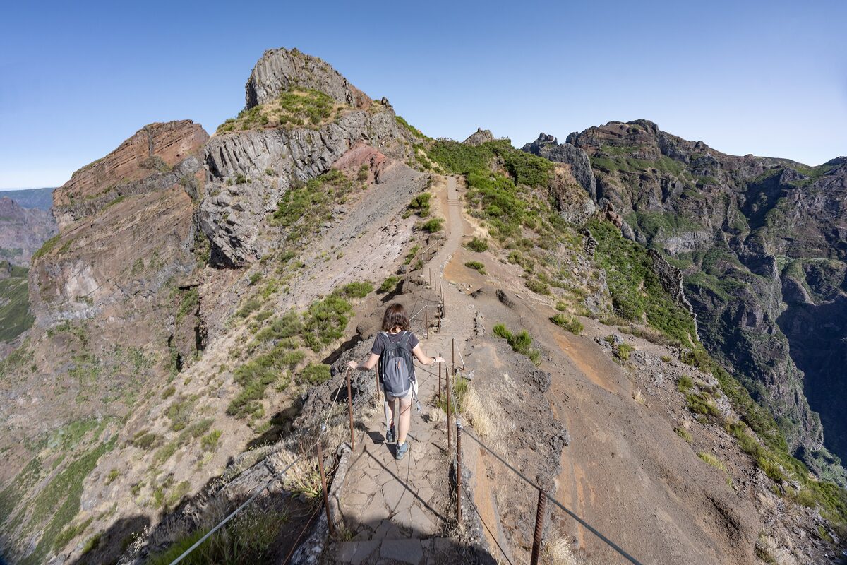 A hiker on the rocky PR1 path along the ridge, with a steep drop and a rope railing beside the trail