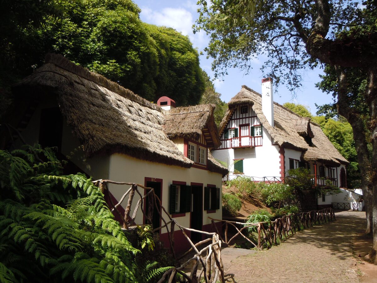 Two thatched, white-walled cottages with dark shutters among ferns and trees at Queimadas Forest Park, with a rustic wooden fence along a gravel path
