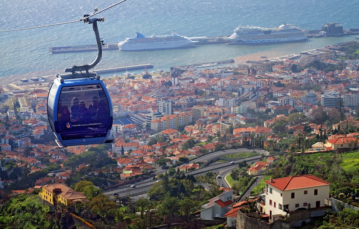A blue cable car gondola high above Funchal's red roofs, with two cruise ships moored in the harbor behind