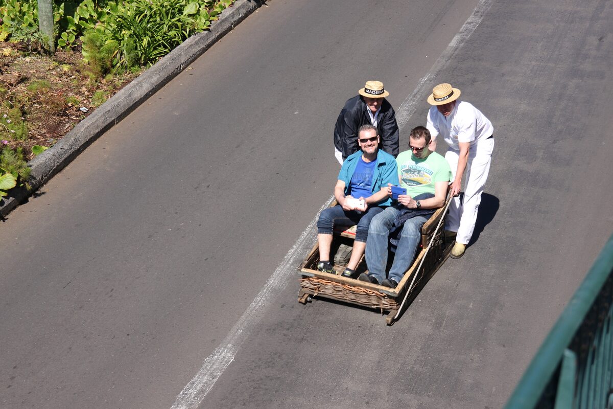 A wicker toboggan with two passengers sliding down a steep Monte road, steered by two men in straw hats and white trousers