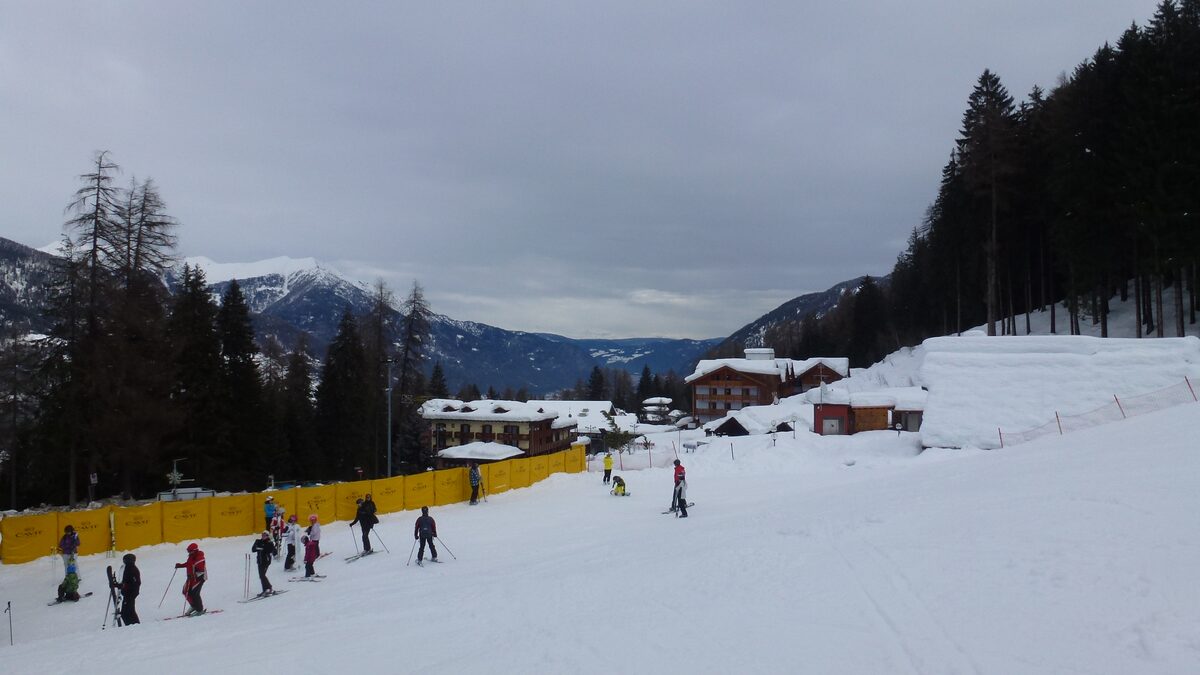 Skiers on a snowy slope beside the hotels of Folgarida, under an overcast winter sky
