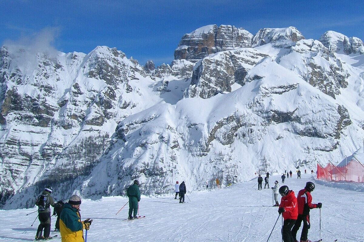Skiers on a sunny snow slope below the snow-covered rock peaks of the Brenta Dolomites