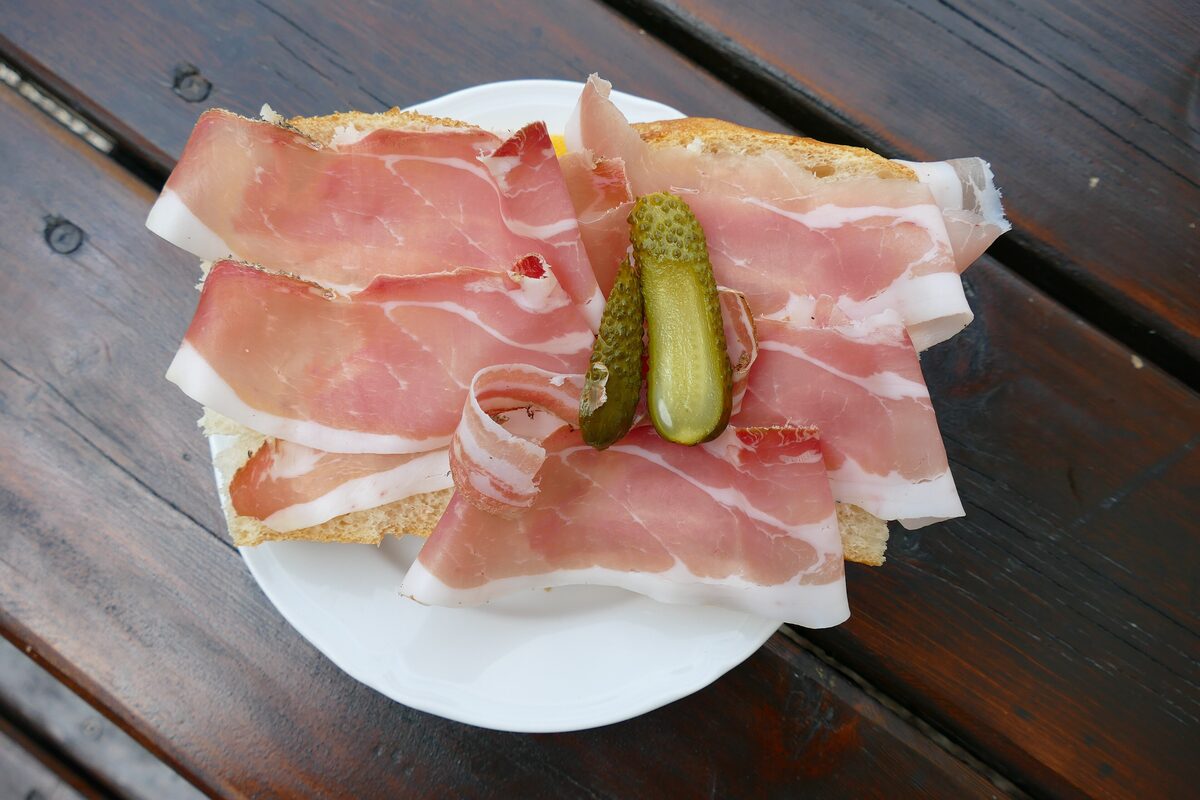 Thin slices of speck on a bread roll with two pickles, on a wooden table