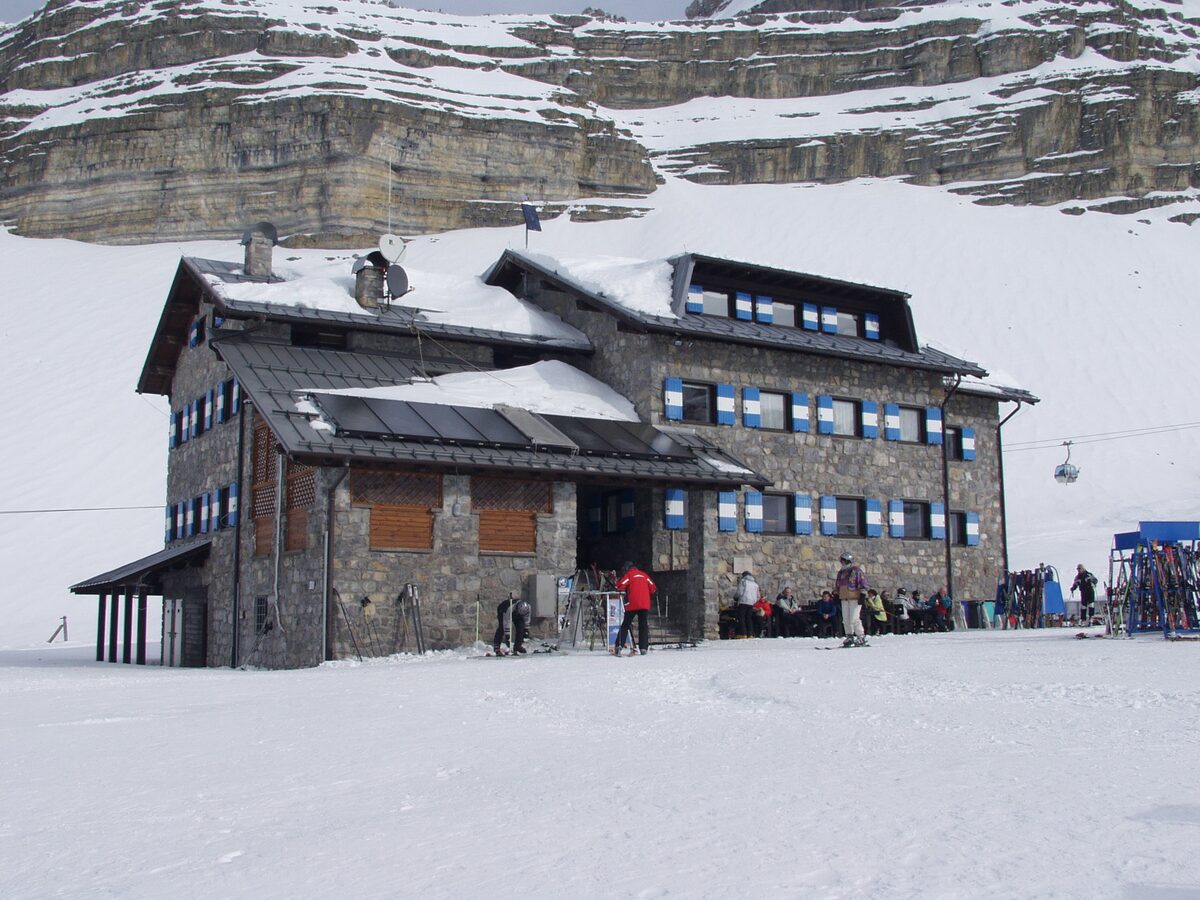 Rifugio Graffer, a stone mountain hut with blue shutters, in deep snow below a rock wall, with a chairlift behind