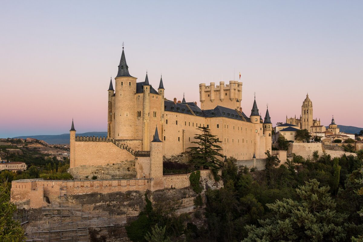 The towers and walls of the Alcázar of Segovia on its rocky spur, with a pink evening sky behind