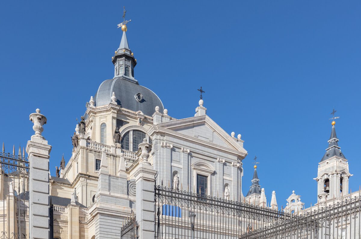 The dome and bell towers of Almudena Cathedral against a blue sky, behind an iron fence