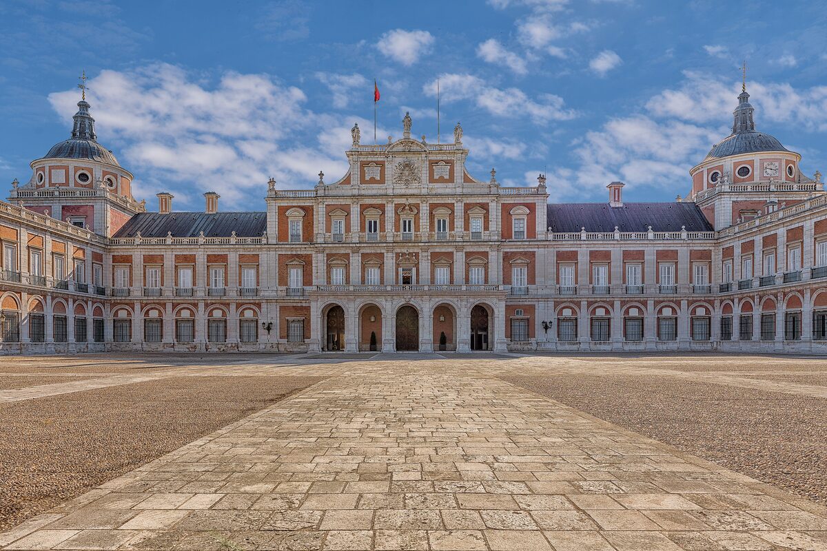 The pink and cream Royal Palace of Aranjuez seen across its stone courtyard under a blue sky with clouds