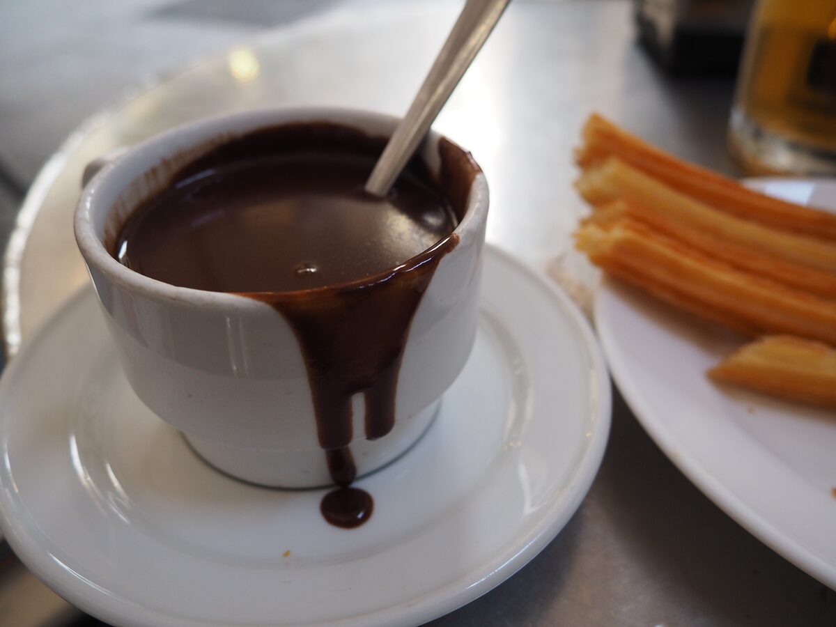 A cup of thick hot chocolate dripping over the rim, with churros on a plate beside it, at Chocolatería San Ginés