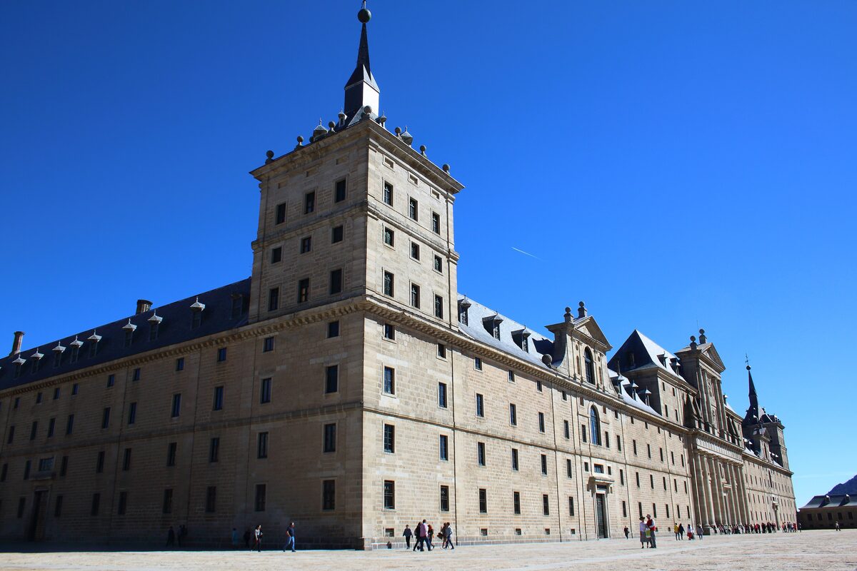 The granite corner tower and long facade of the El Escorial monastery under a deep blue sky, with a few visitors on the square