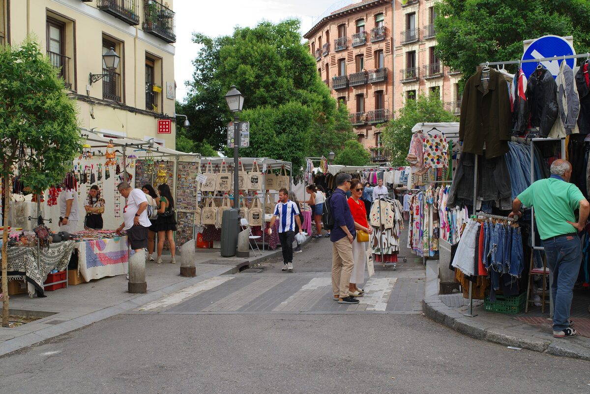 Clothing and bric-a-brac stalls lining a street at the Rastro flea market, with shoppers browsing