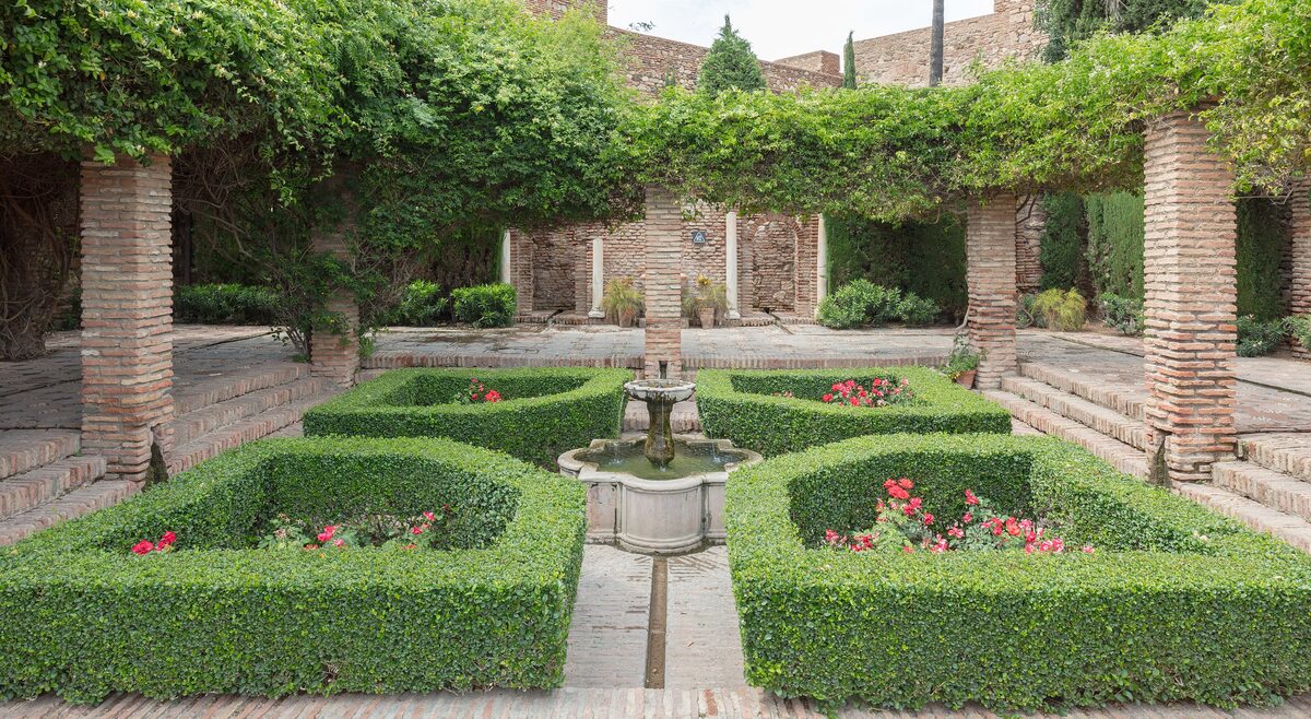A garden courtyard in the Alcazaba with clipped box hedges, a stone fountain and a vine pergola on brick pillars