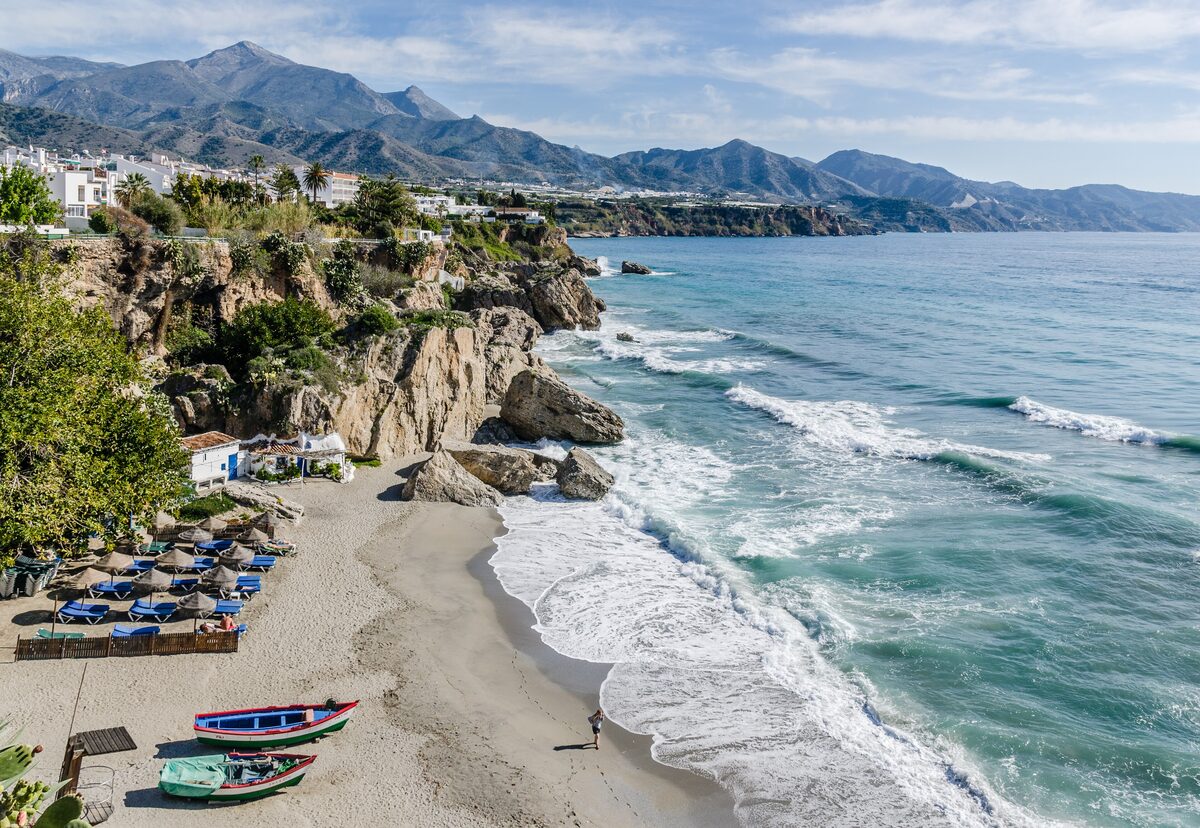 The view from the Balcón de Europa in Nerja: a sandy cove with boats and surf below a rocky headland, mountains beyond