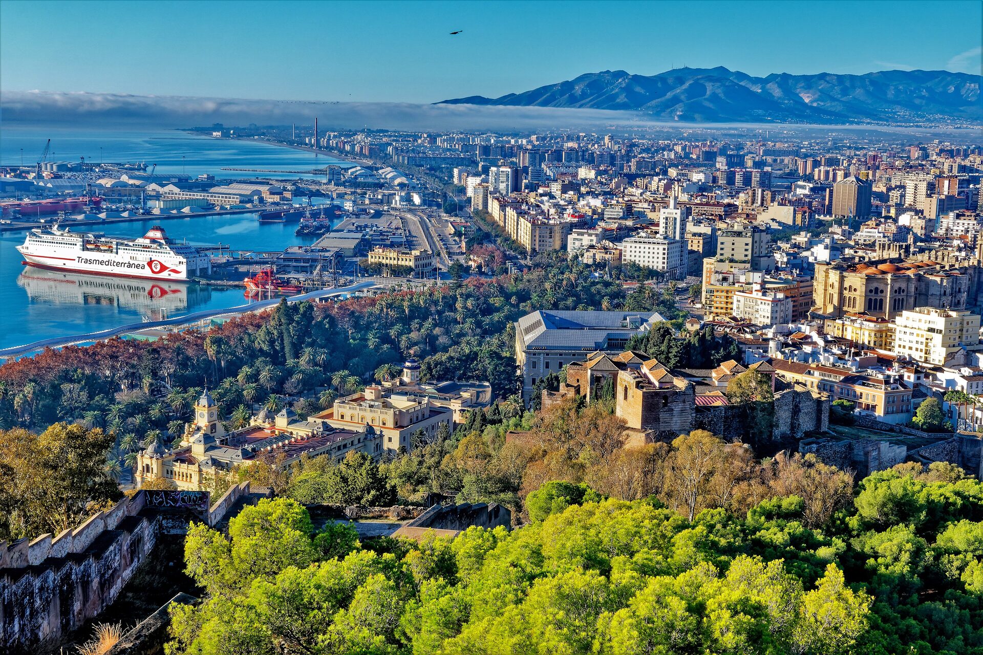 Málaga from the castle walls: the Alcazaba among pines, cathedral roofs, a ferry in the port and mountains behind