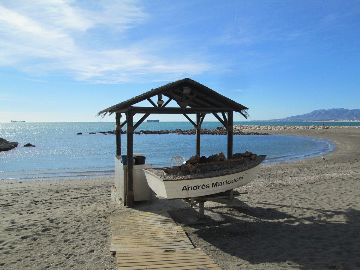 A boat full of firewood under a thatched shelter on Pedregalejo beach, the espeto grill setup, with calm sea beyond