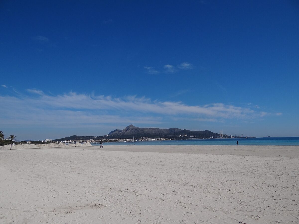 A wide pale-sand beach on the Bay of Alcúdia under a deep blue sky, with the headland in the distance