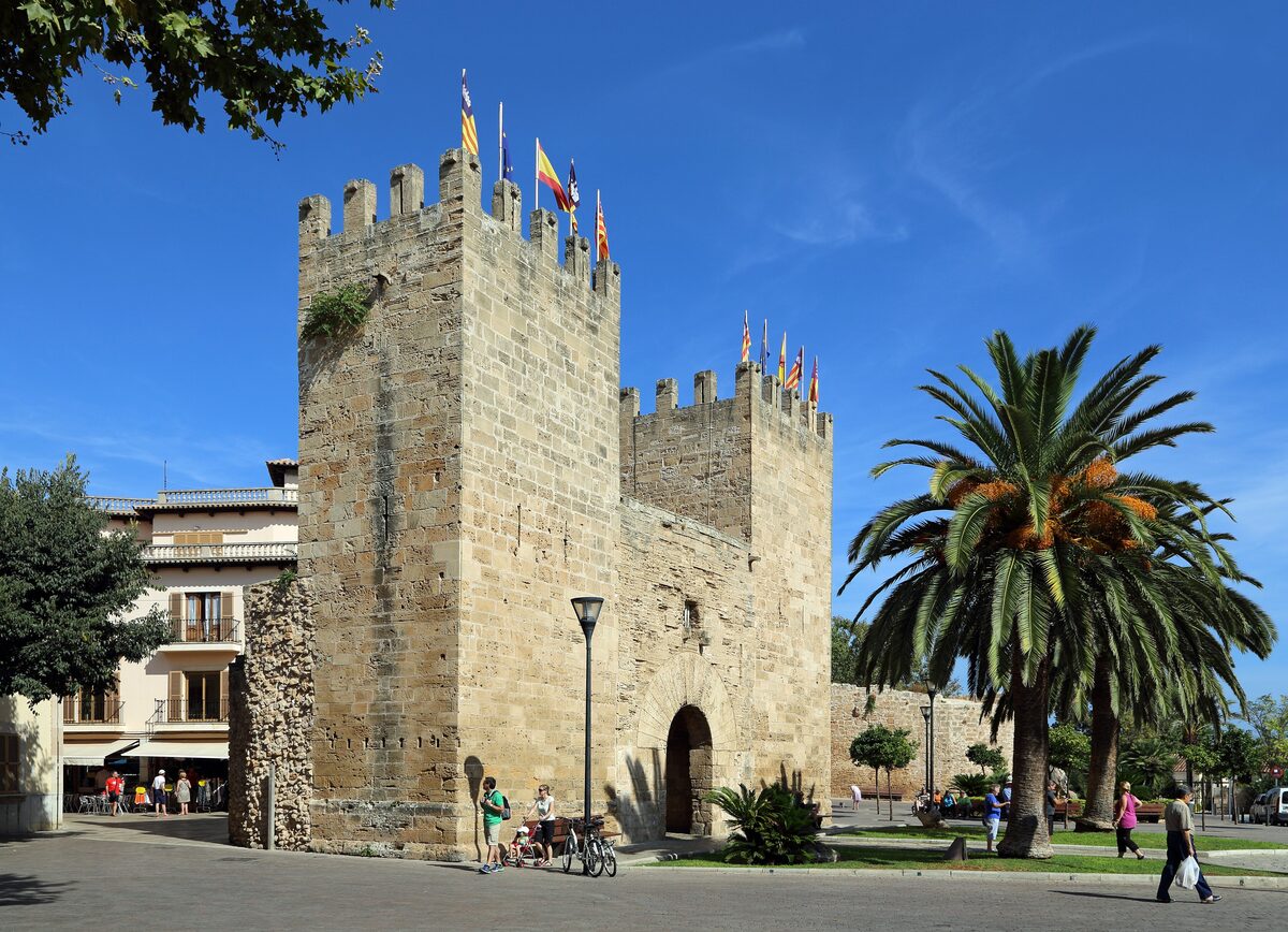 The battlemented towers of the Porta del Moll, a gate in the medieval walls of Alcúdia, with a palm tree beside it