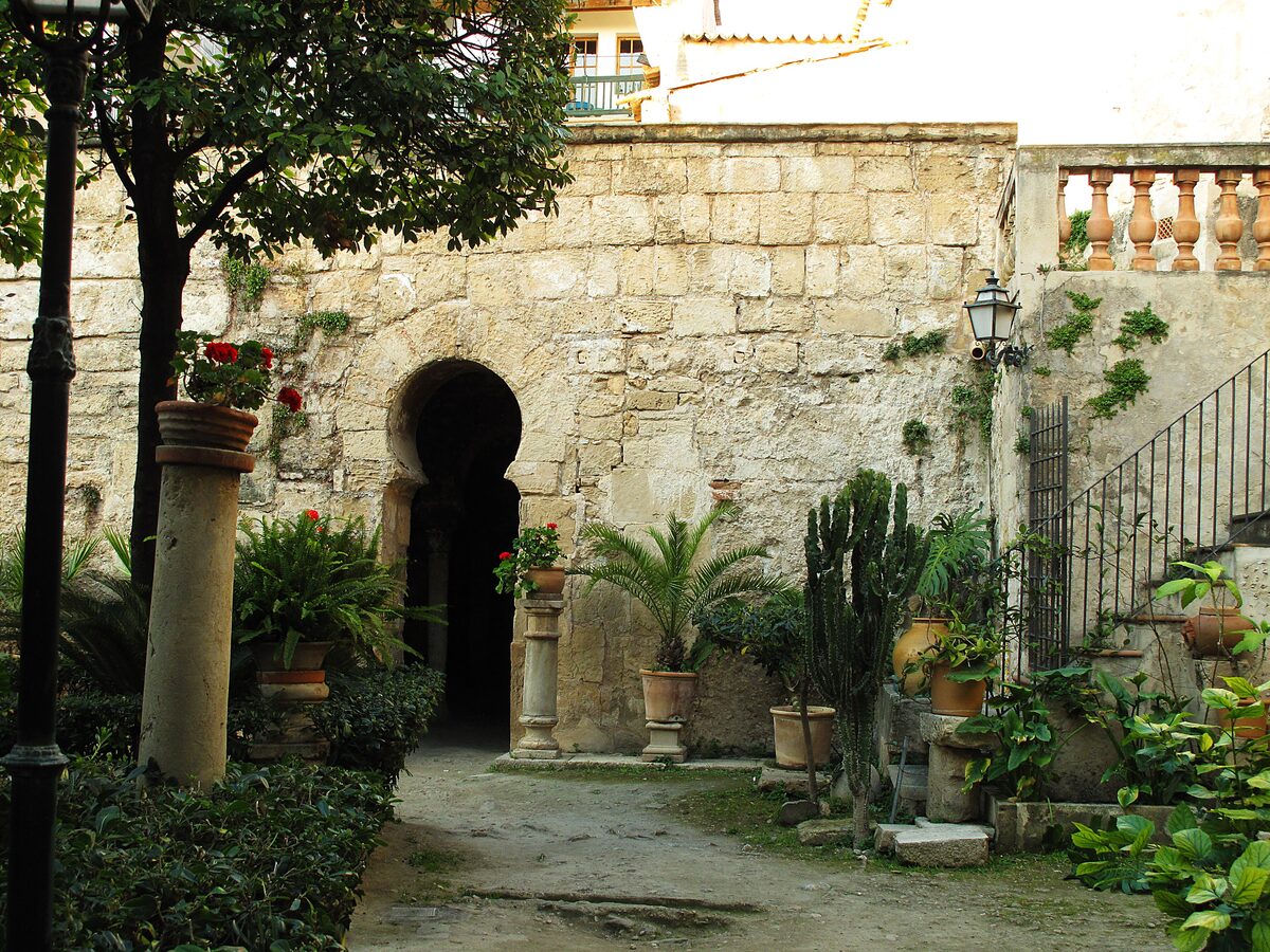 The courtyard of the Arab Baths, a horseshoe-arched doorway in an old stone wall with potted plants and a staircase