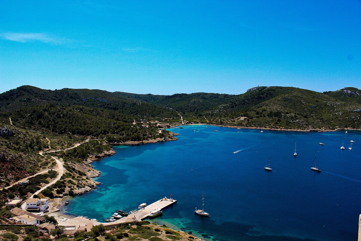 Aerial view of a blue bay on Cabrera with boats moored at a small jetty and pine-covered hills around the water