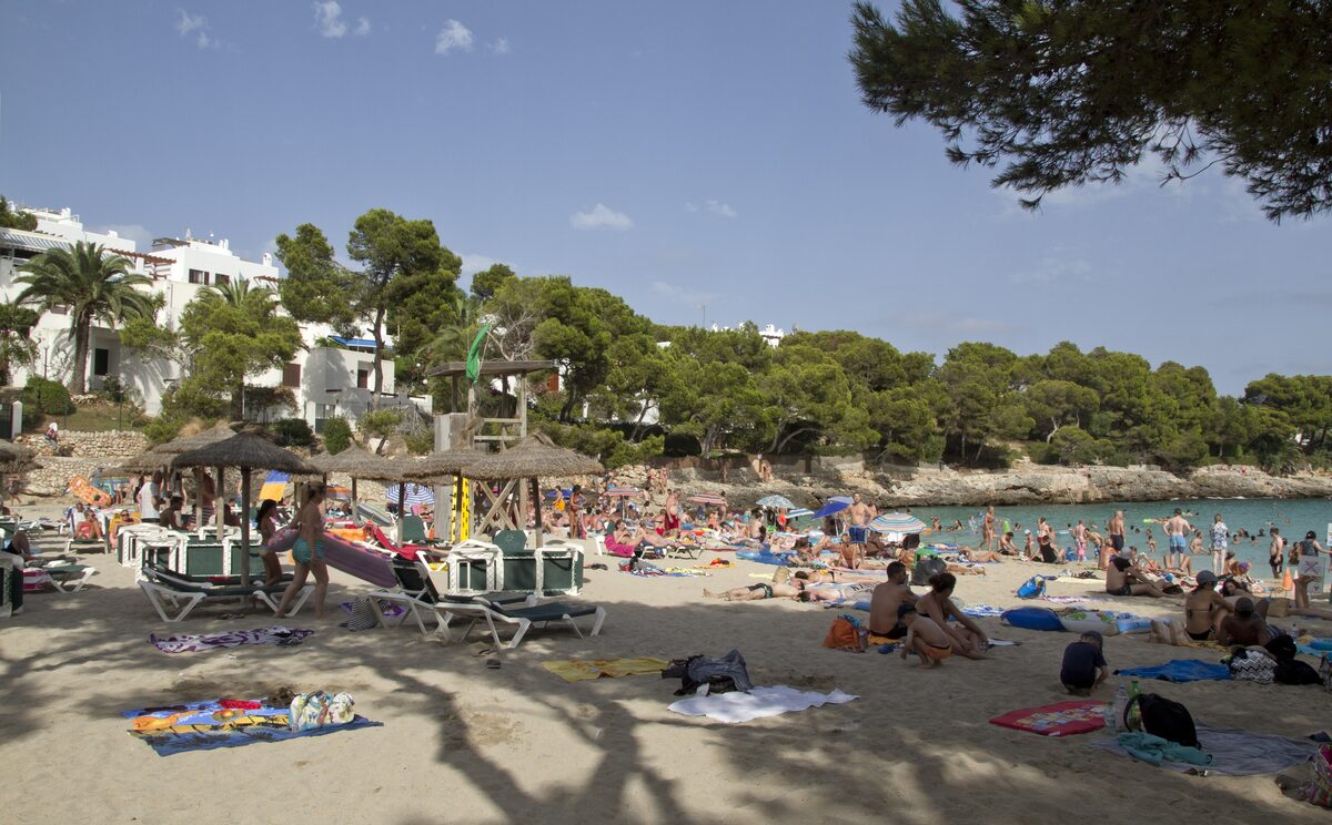 Sunbathers, parasols and sun loungers on the sand of a Cala d'Or beach, with pine trees and white houses around the bay