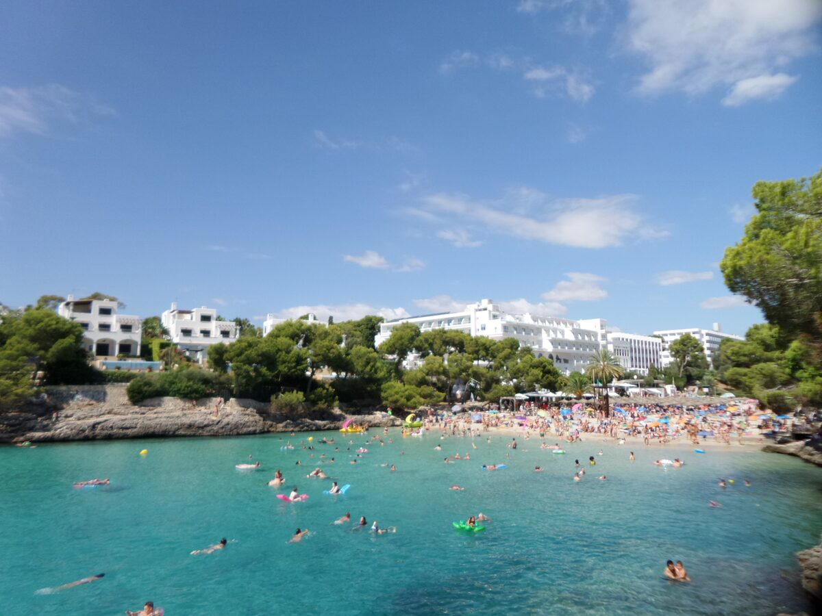 Cala Gran in Cala d'Or on a sunny day: turquoise water full of swimmers, a sandy shore and white hotels among pines