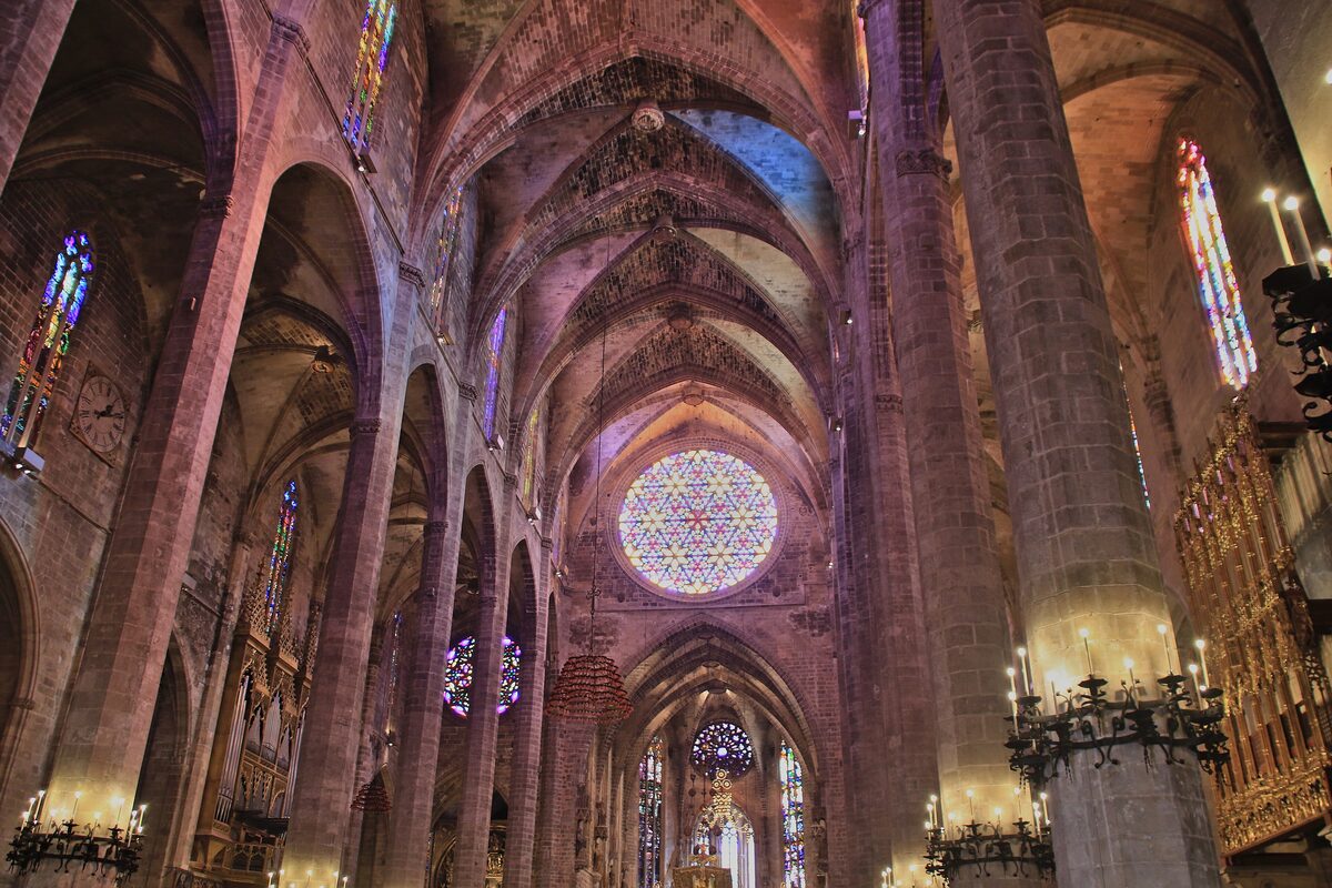 The inside of Palma Cathedral, tall Gothic pillars and ribbed vaults under a large rose window, with stained glass and candles in the nave