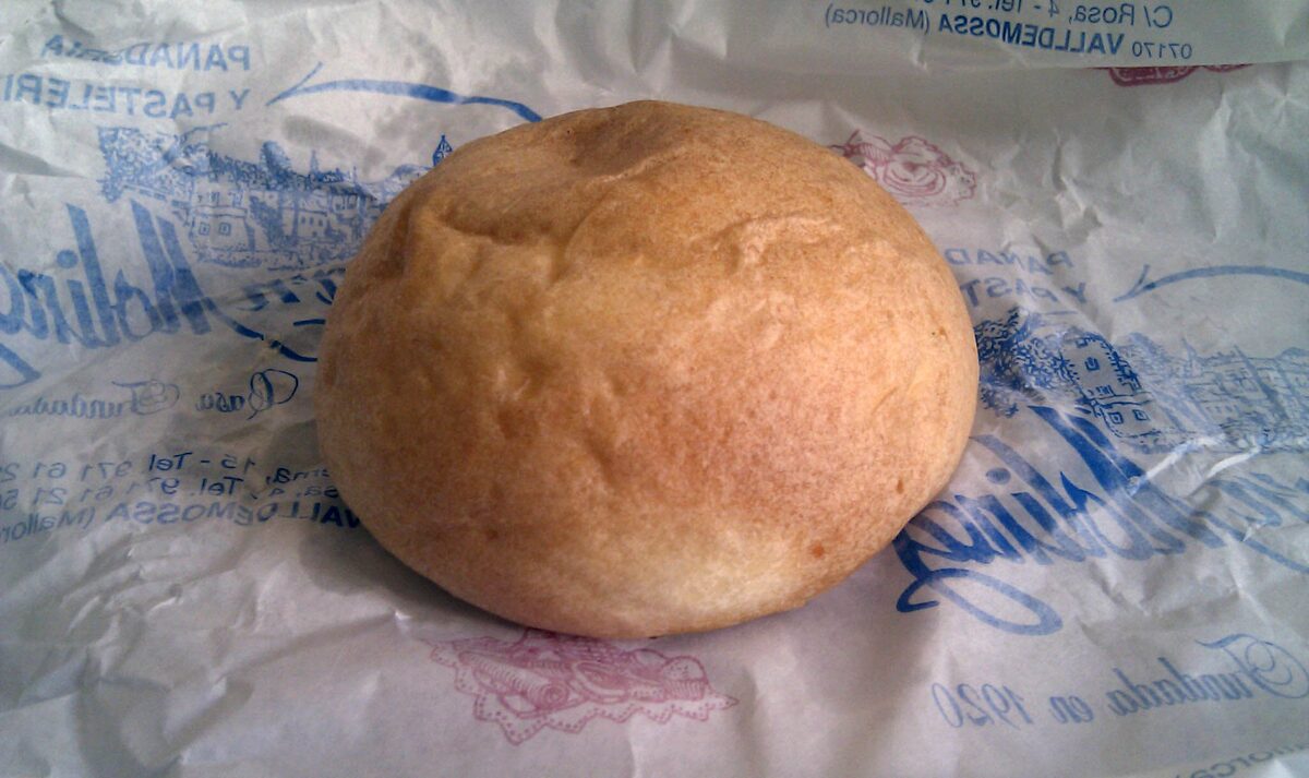 A round potato coca bun, golden and soft, on a sheet of bakery paper printed with the name of a Valldemossa bakery