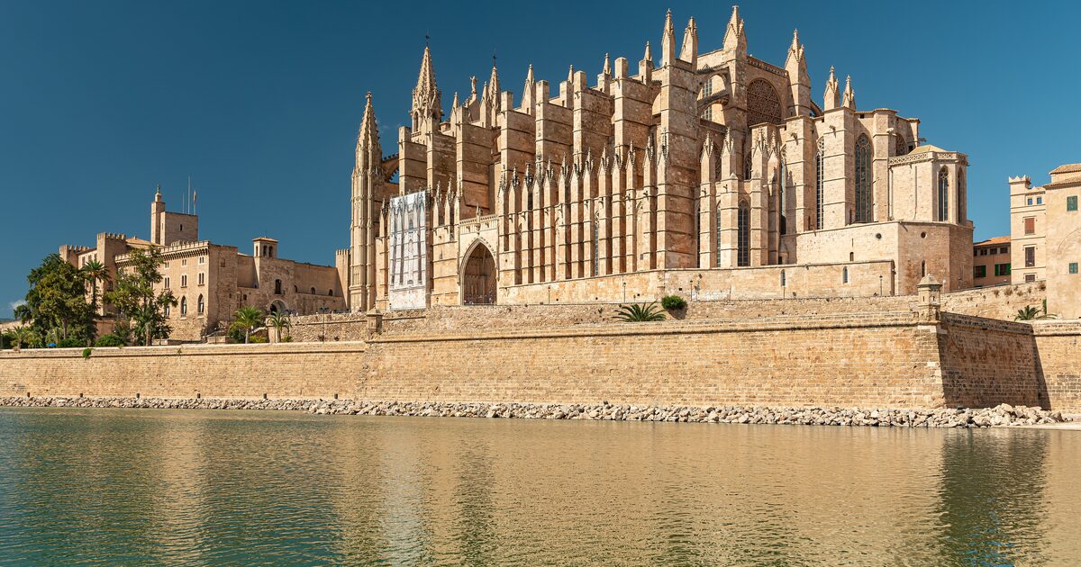 Palma Cathedral, a sandstone Gothic church with a forest of pinnacles, rising behind the sea wall and water of the Parc de la Mar under a clear blue sky