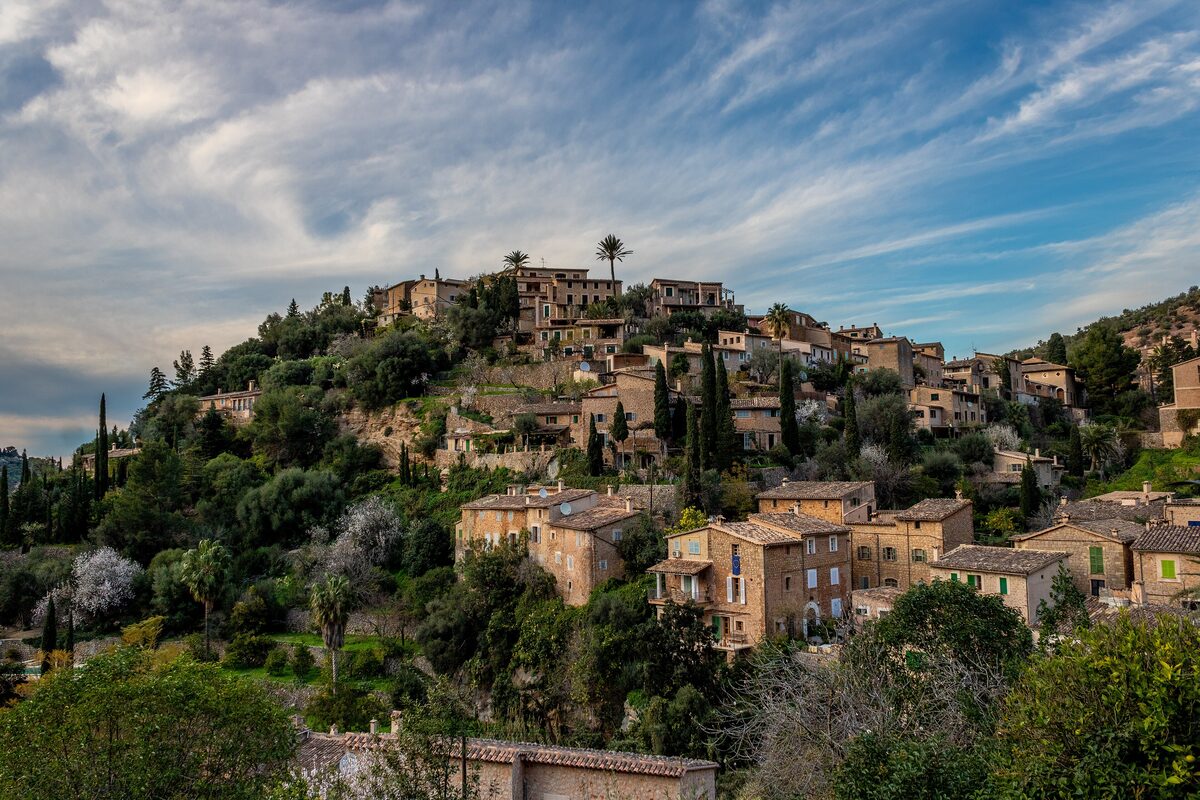 Deià, a village of stone houses stacked on a steep green hillside under a sky with thin clouds