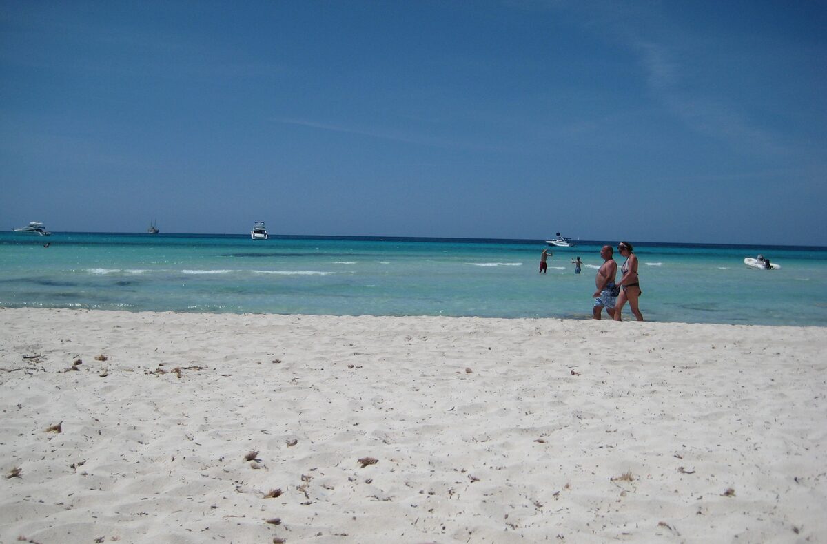 Es Trenc's white sand in the foreground and turquoise water beyond, with a couple walking at the shore