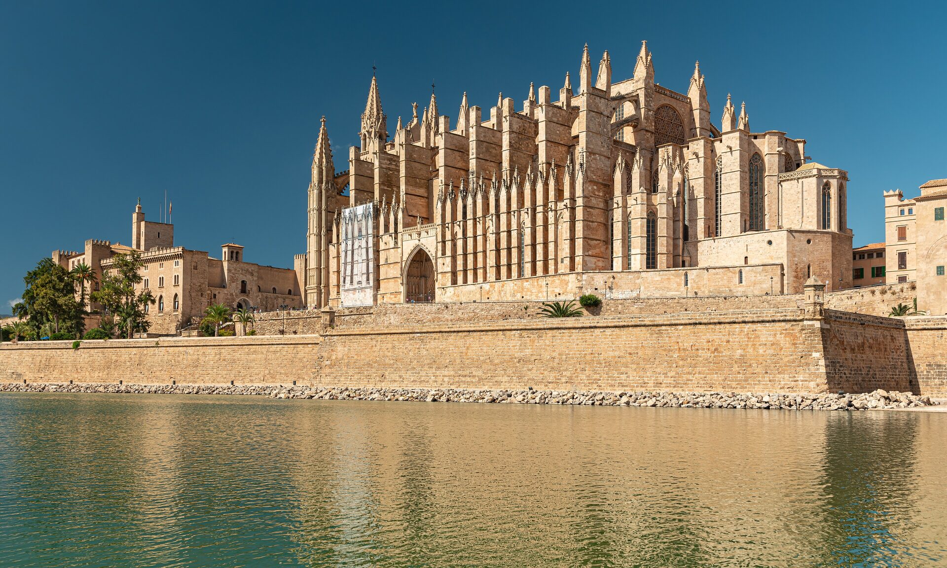 Palma Cathedral, a sandstone Gothic church with a forest of pinnacles, rising behind the sea wall and water of the Parc de la Mar under a clear blue sky