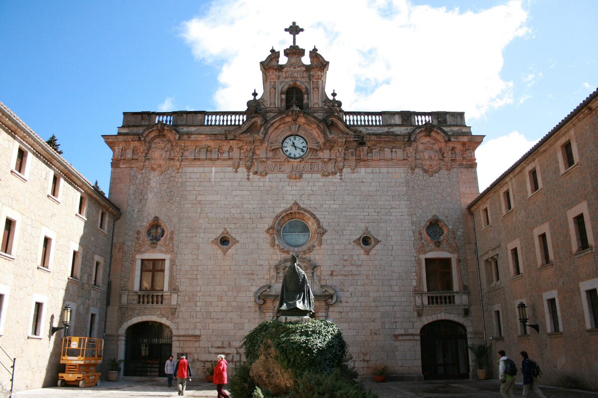 The front of the Santuari de Lluc basilica, a stone monastery with a clock tower and a statue in the courtyard