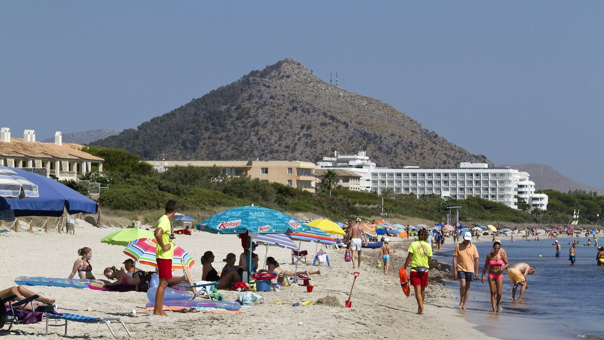 Platja de Muro on a sunny day: pale sand, beach umbrellas, shallow water and a hill behind the hotels