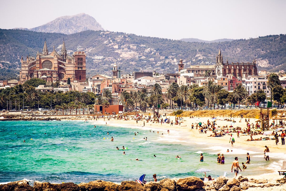 Palma's city beach on a sunny day, with swimmers in turquoise water and the cathedral rising above the town behind