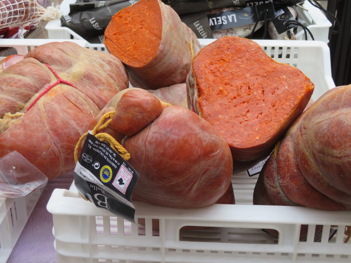 Whole and sliced sobrassada sausages, soft and orange-red, in crates at a market stall