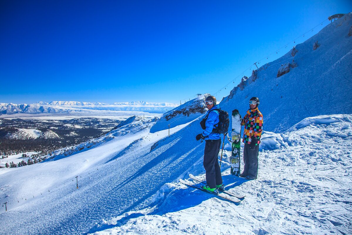 Two skiers on the rim of a steep, sunlit summit bowl