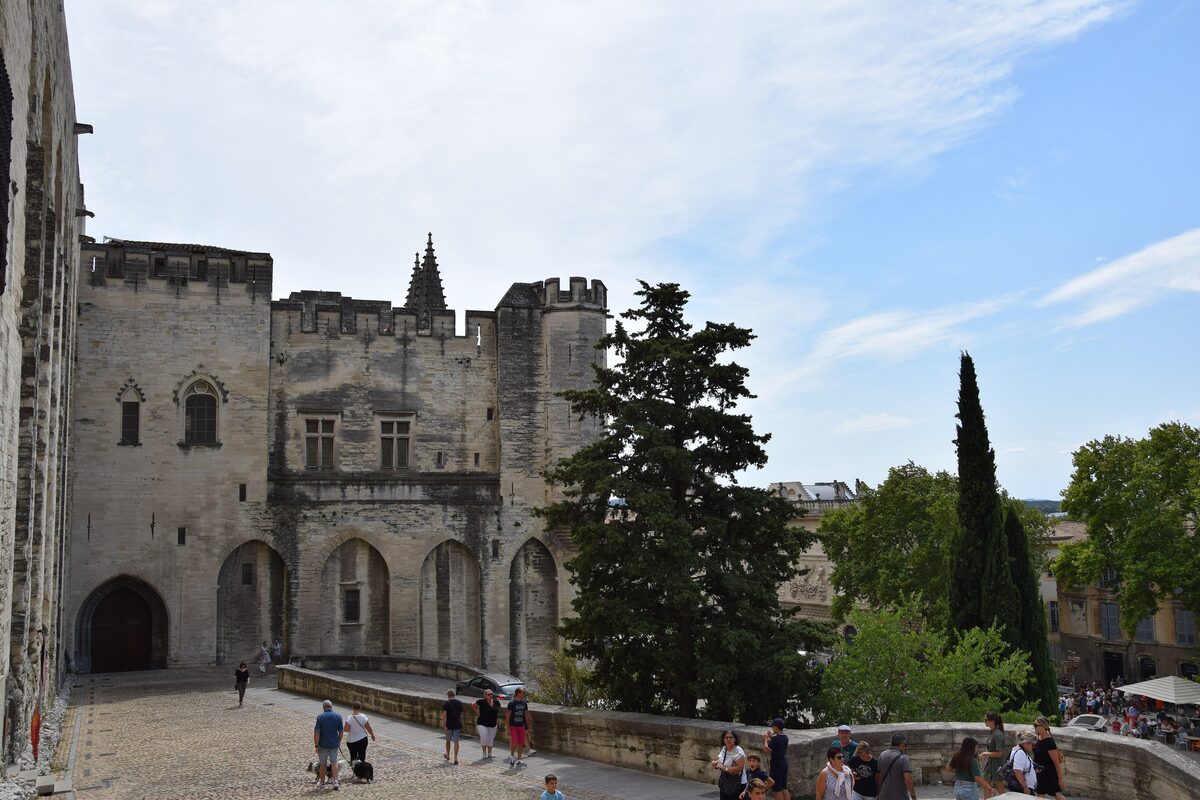 A fortified wing of the Palais des Papes in Avignon, with crenellated towers above the cobbled forecourt and visitors on the terrace