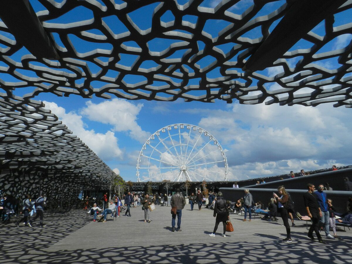The black lattice shell of the MuCEM on its esplanade, with visitors walking beneath it and the Marseille Ferris wheel behind