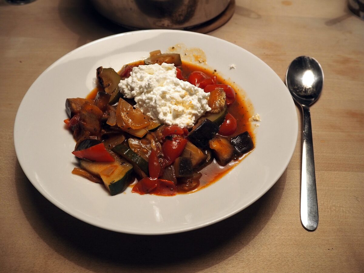 A plate of ratatouille with aubergine, courgette and tomato, topped with a spoonful of soft white cheese