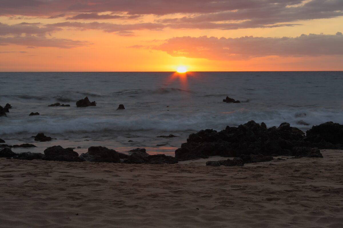 The sun setting over the sea at Keawakapu Beach, with dark lava rocks on the shore